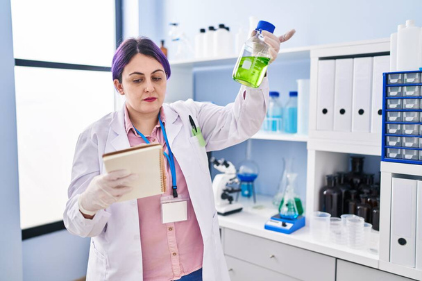 Jeune belle plus taille femme scientifique lecture ordinateur portable tenant bouteille avec du liquide au laboratoire - Photo, image