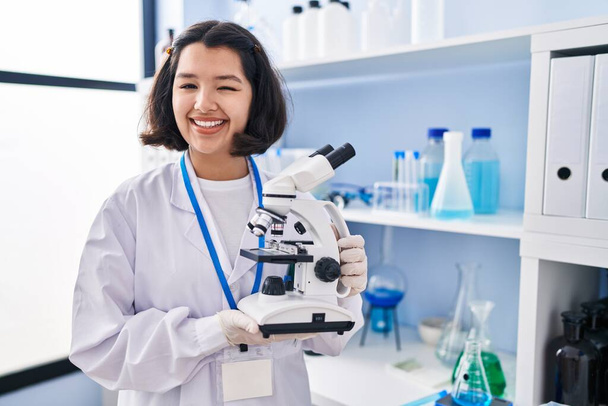 Mladá hispánka pracující v laboratoři vědců, která drží mikroskop a mrká na kameru se sexy výrazem, veselou a šťastnou tváří.  - Fotografie, Obrázek