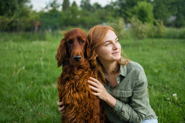 Piękna kobieta przytulająca i całująca irlandzkiego psa setera. Pies i właściciel razem na zewnątrz. Miłość i przyjaźń między psem a właścicielem w parku. prawdziwi przyjaciele na zawsze, ludzie zwierzęta koncepcja - Zdjęcie, obraz