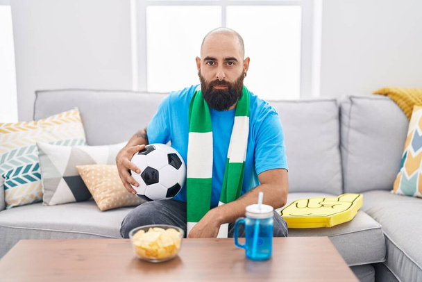 Молодой латиноамериканец с бородой и татуировками футбольный хулиган держит мяч, поддерживая командное мышление и трезвое выражение лица, выглядящее уверенным в себе  - Фото, изображение