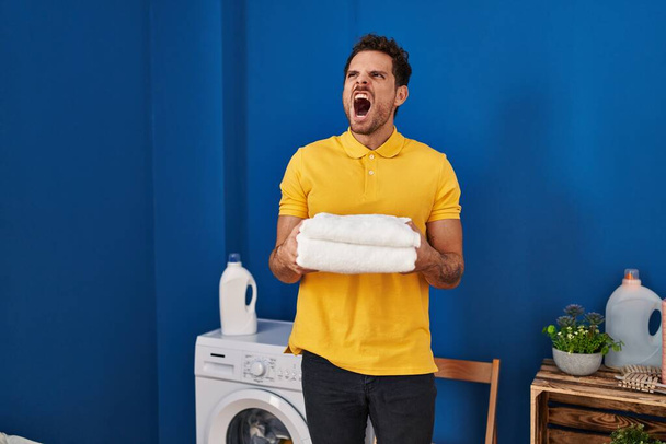 Young hispanic man holding clean towels at laundry room angry and mad screaming frustrated and furious, shouting with anger. rage and aggressive concept.  - 写真・画像
