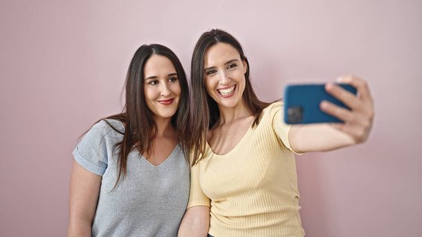Zwei selbstbewusst lächelnde Frauen machen Selfie per Smartphone vor isoliertem rosa Hintergrund - Foto, Bild