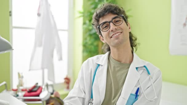 Jeune homme hispanique médecin souriant confiant tenant bouteille de médicaments à la clinique - Séquence, vidéo