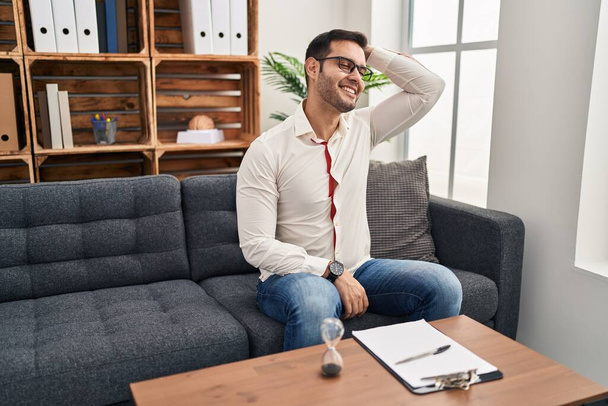 Νεαρός ισπανόφωνος άνδρας με γενειάδα εργάζεται στο γραφείο διαβούλευσης χαμογελώντας αυτοπεποίθηση αγγίζοντας τα μαλλιά με το χέρι προς τα πάνω χειρονομία, θέτοντας ελκυστική και μοντέρνα  - Φωτογραφία, εικόνα