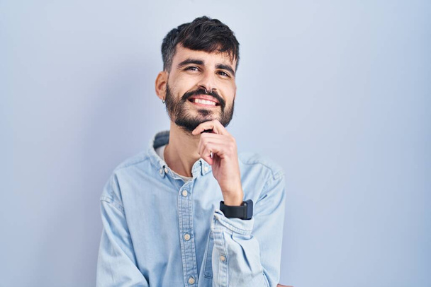 Молодий іспаномовний чоловік з бородою, що стоїть над синім тлом з рукою на підборідді думаючи про питання, стомлюючий вираз. усміхнене і вдумливе обличчя. концепція сумніву
.  - Фото, зображення