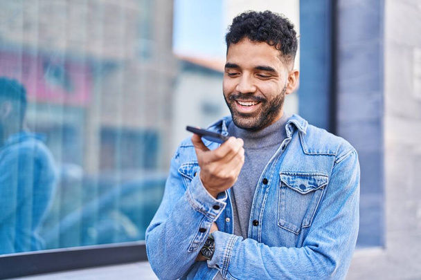 Молодой латиноамериканец уверенно улыбается, разговаривая по смартфону на улице - Фото, изображение