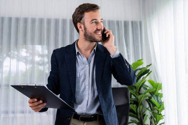 Ciężko pracujący biznesmen stoi pewnie w nowoczesnym biurze, wykonując przekonywujący telefon do klienta. Pracownik biurowy rozmawia przez telefon koordynować i zarządzać pracą biznesową z kolegami. Podmiot - Zdjęcie, obraz