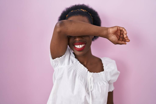 Mujer africana con el pelo rizado de pie sobre fondo rosa cubriendo los ojos con el brazo sonriendo alegre y divertido. concepto ciego.  - Foto, imagen