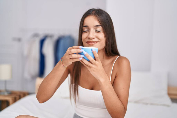Νεαρή όμορφη ισπανίδα γυναίκα πίνει καφέ καθισμένη στο κρεβάτι στο υπνοδωμάτιο - Φωτογραφία, εικόνα