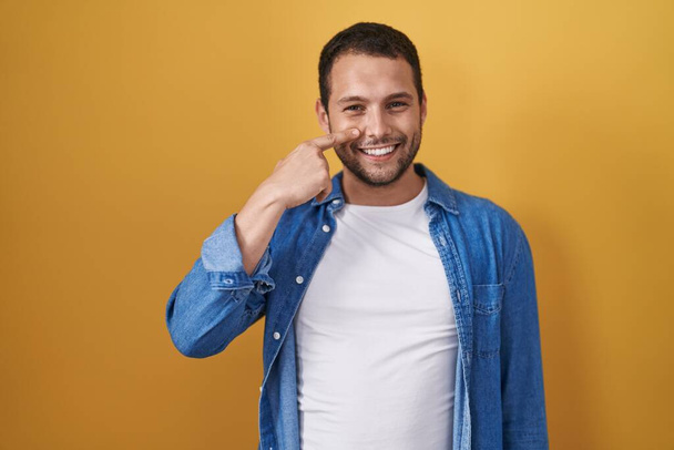 Латиноамериканец, стоящий на жёлтом фоне, указывая пальцем руки на лицо и нос, весело улыбаясь. концепция красоты  - Фото, изображение