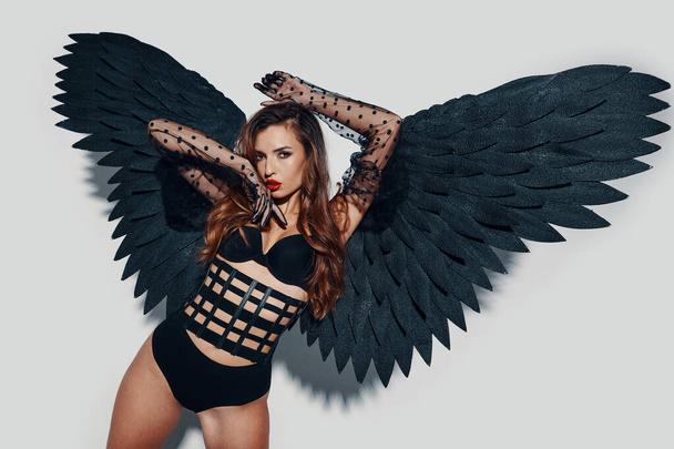 Jovem atraente em lingerie vestindo asas de anjo preto contra fundo cinza - Foto, Imagem