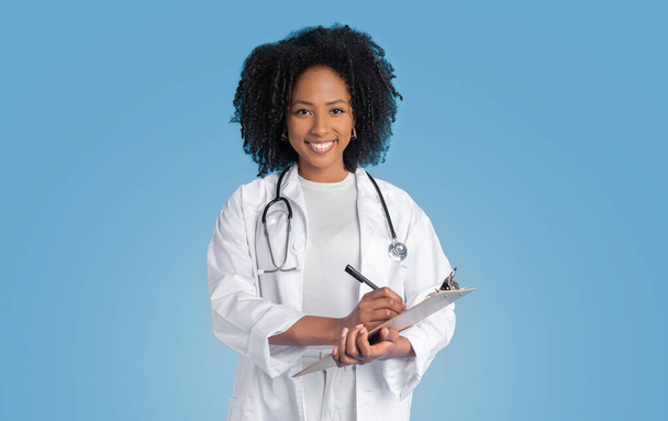 Mosolygó fiatal afro-amerikai göndör nő orvos terapeuta fehér kabátban jegyzetel tabletta, elszigetelt kék háttér, stúdió. Orvosi szolgálat és vizsga, egészségügyi ellátás és kezelés - Fotó, kép