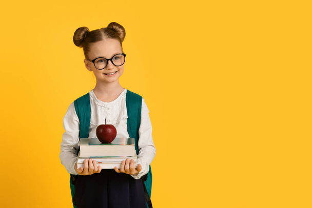 Education Concept. Little Cute Schoolgirl Holding Stack Of Books With Red Apple On Top, Smiling Nerdy Female Child With Backpack Posing Isolated Over Yellow Studio Background, Copy Space - Φωτογραφία, εικόνα