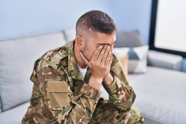 Young hispanic man army soldier sitting on sofa stressed at home - Foto, imagen