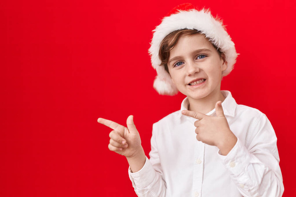 Little caucasian boy wearing christmas hat over red background smiling and looking at the camera pointing with two hands and fingers to the side.  - Фото, изображение