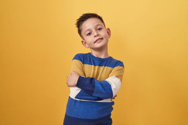 Kleine Latijns-Amerikaanse jongen die over een gele achtergrond staat vrolijk gezicht glimlachend met gekruiste armen kijkend naar de camera. positieve persoon.  - Foto, afbeelding
