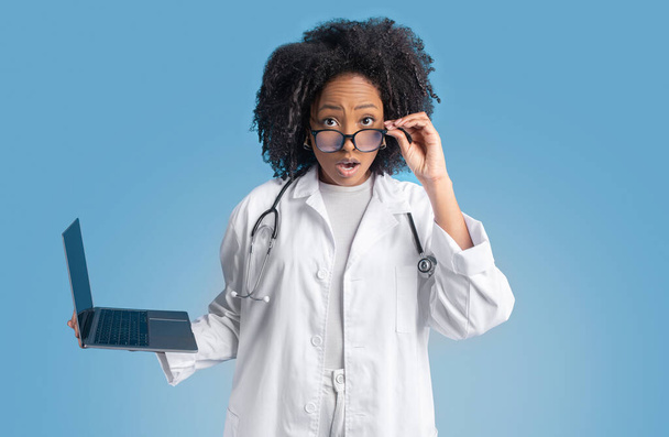 Σοκαρισμένη νεαρή αφροαμερικανίδα σγουρή θεραπεύτρια γιατρός με λευκό παλτό με φορητό υπολογιστή βγάζει γυαλιά, απομονωμένη σε μπλε φόντο, στούντιο. Έκπληξη, καλά νέα, ιατρική υπηρεσία, υγειονομική περίθαλψη - Φωτογραφία, εικόνα