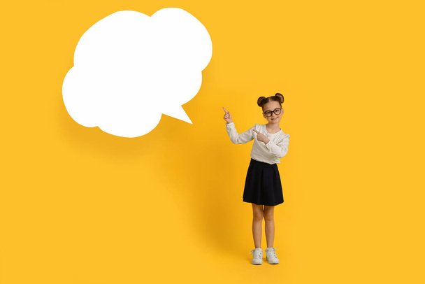 Smiling Little Schoolgirl Pointing Aside At Empty Speech Bubble, Cute Preteen Female Child Demonstrating Copy Space For Educational Offer While Standing Over Yellow Studio Background, Full Length - Φωτογραφία, εικόνα