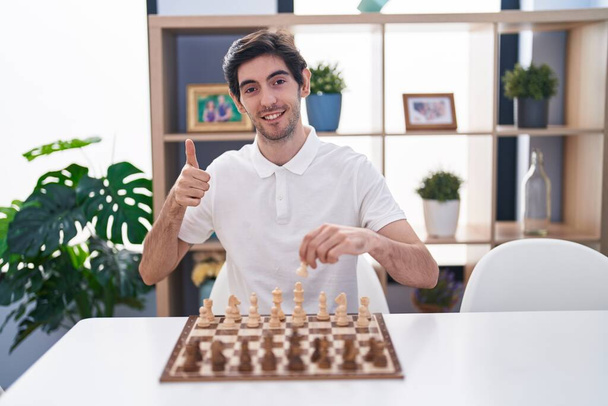 Masada oturmuş satranç oynayan genç İspanyol adam mutlu ve olumlu gülümsüyor, başparmağıyla mükemmel ve onay işareti yapıyor.  - Fotoğraf, Görsel