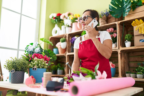 Young beautiful hispanic woman florist talking on smartphone writing on notebook at flower shop - Photo, Image