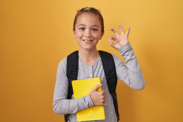 Küçük beyaz çocuk, öğrenci çantası takıyor ve elinde kitap tutuyor. Parmaklarıyla imza atıyor, gülümsüyor ve mükemmel bir sembol.  - Fotoğraf, Görsel