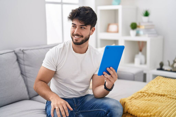 Homem hispânico com barba usando touchpad sentado no sofá olhando para o lado com sorriso no rosto, expressão natural. rindo confiante.  - Foto, Imagem