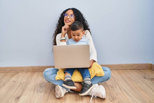 若いヒスパニック系の母親と子供のコンピュータのラップトップの床に手であごの質問については、集中的な表現を考えて座って使用しています。笑顔で思いやりのある顔。疑わしい概念.  - 写真・画像