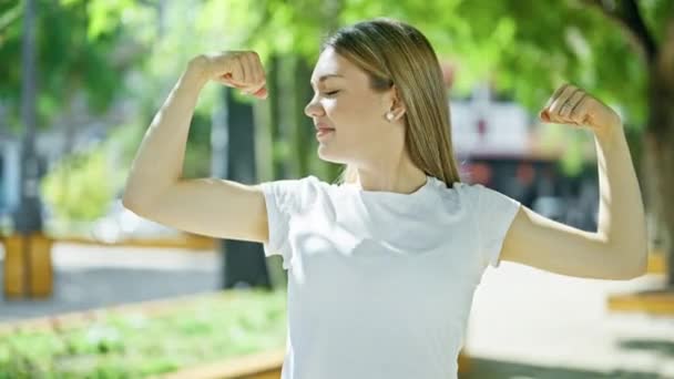 Fiatal szőke nő mosolyog magabiztos csinál erős gesztus karokkal a parkban - Felvétel, videó