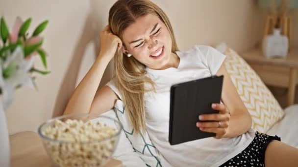 Nuori blondi nainen katsomassa elokuvaa istuu sängyllä syö popcornia makuuhuoneessa - Materiaali, video