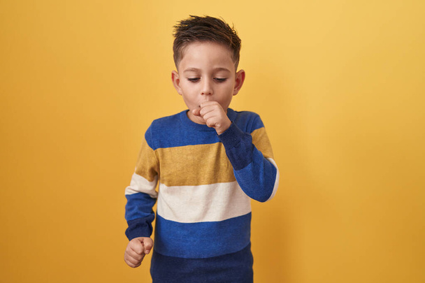 黄色の背景に立っている小さなヒスパニック系の少年は具合が悪く感じ、風邪や気管支炎の症状として咳をします。ヘルスケアの概念.  - 写真・画像