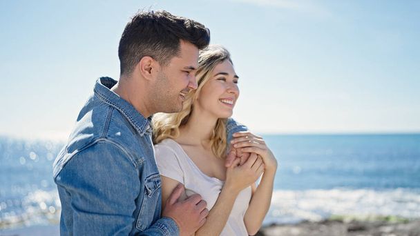 Мужчина и женщина улыбаются уверенно обнимая друг друга на берегу моря - Фото, изображение