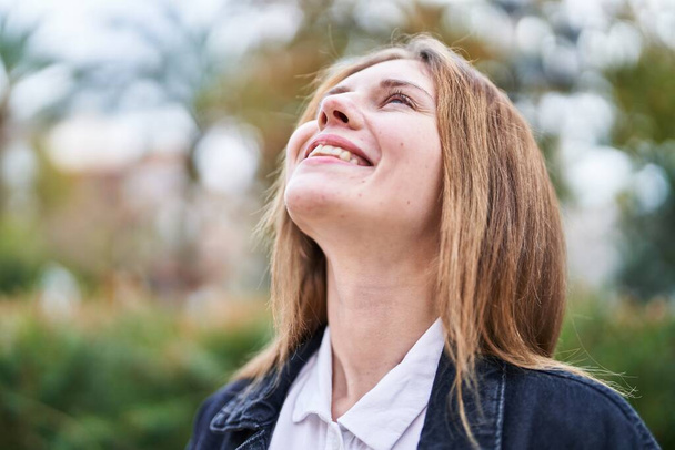 Νεαρή ξανθιά γυναίκα χαμογελά με αυτοπεποίθηση κοιτάζοντας στον ουρανό στο πάρκο - Φωτογραφία, εικόνα