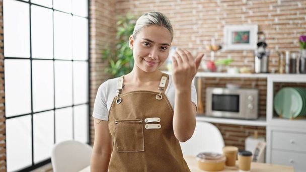 Young beautiful hispanic woman wearing apron doing coming gesture at dinning room - Foto, afbeelding