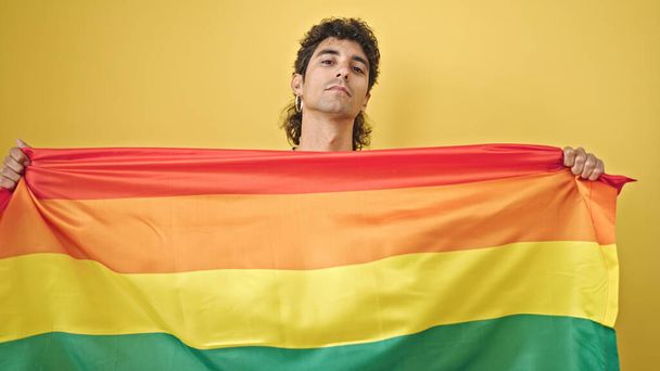 Joven hispano sosteniendo bandera de arco iris con expresión relajada sobre fondo amarillo aislado - Foto, imagen