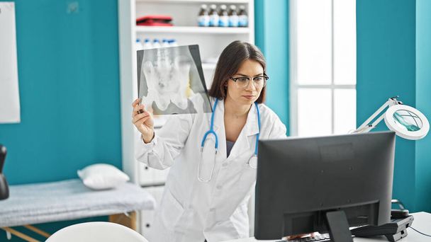 Jovem bela mulher hispânica médico olhando para xray usando computador de pé na clínica - Foto, Imagem