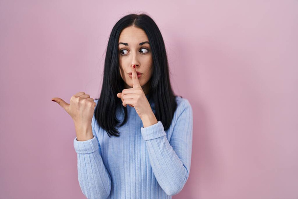 Hispanic woman standing over pink background asking to be quiet with finger on lips pointing with hand to the side. silence and secret concept.  - Фото, зображення