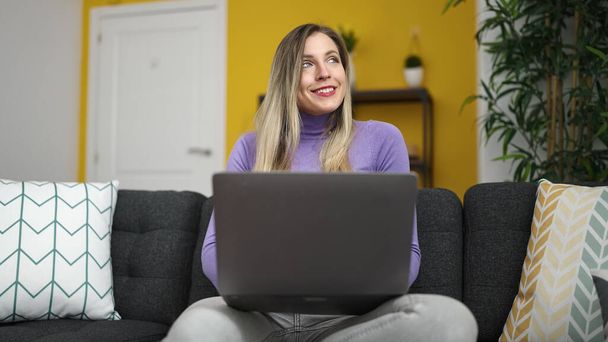 Mujer rubia joven usando el ordenador portátil sentado en el sofá en casa - Foto, imagen