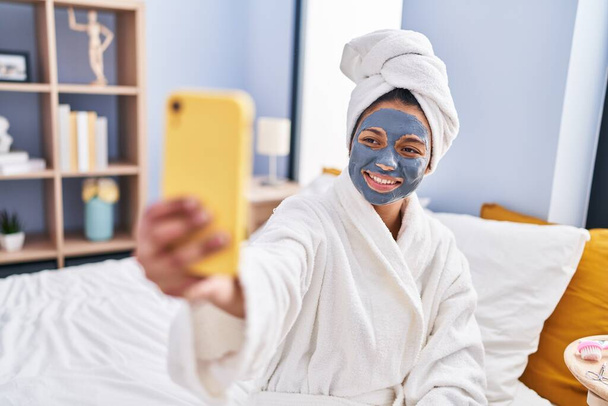 Молодая латиноамериканка, надевающая кремовую маску, делает селфи на смартфоне в спальне - Фото, изображение