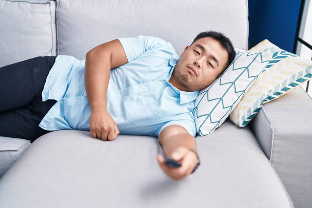 Joven hombre chino viendo la televisión tumbado en el sofá con expresión aburrida en casa - Foto, imagen