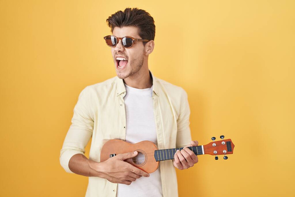 Young hispanic man playing ukulele over yellow background angry and mad screaming frustrated and furious, shouting with anger. rage and aggressive concept.  - Fotó, kép