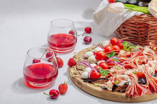Picnic food concept. Basket with wine and baguette, set of snacks on skewers. Meat, cheese, vegetables, fruits and flowers bouquet. Hard light, dark shadows, stone background, copy space - Foto, Imagen