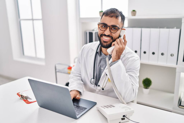 Młody latynoski lekarz korzystający z laptopa rozmawiający przez telefon w klinice - Zdjęcie, obraz
