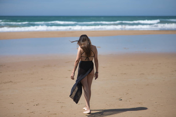 Attractive young woman in black swimsuit and sarong, solitary and quiet, walking on the beach, seen from the back. Concept tranquility, peace, wind, travel, vacation. - Photo, Image