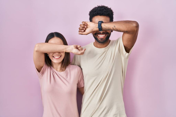 Joven pareja hispana juntos sobre fondo rosa sonriente alegre jugando a echar un vistazo a un boo con las manos mostrando la cara. sorprendido y salido  - Foto, Imagen