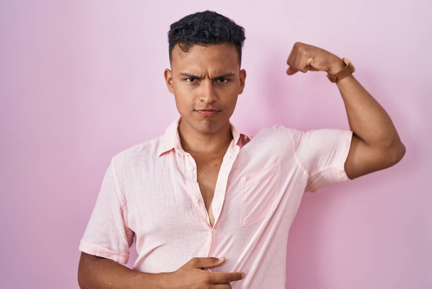 Mladý hispánec muž stojící nad růžovým pozadím silná osoba ukazující sval paže, sebevědomý a hrdý na moc  - Fotografie, Obrázek