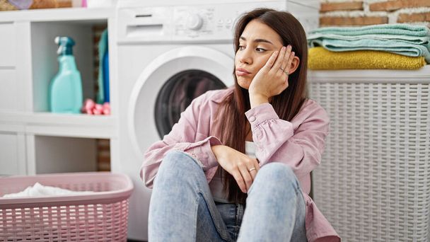 Νέα όμορφη ισπανόφωνη γυναίκα ακουμπά στο πλυντήριο ρούχων με λυπητερή έκφραση στο πλυσταριό - Φωτογραφία, εικόνα
