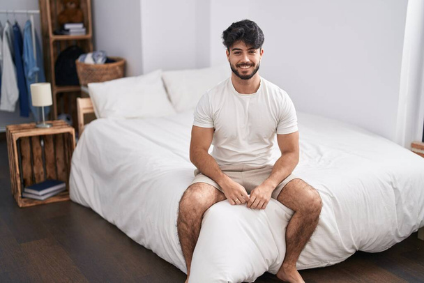 Молодий іспаномовний чоловік посміхається впевнено сидить у ліжку в спальні
 - Фото, зображення