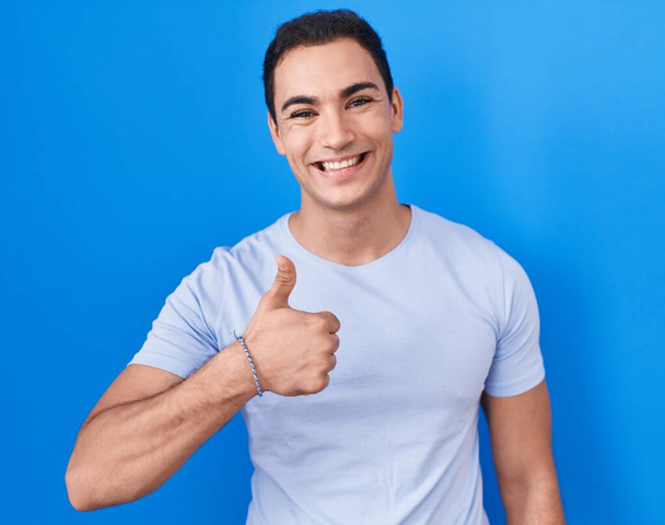 Young hispanic man standing over blue background doing happy thumbs up gesture with hand. approving expression looking at the camera showing success.  - Zdjęcie, obraz