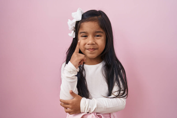 Petite fille hispanique debout sur fond rose souriant avec une idée ou une question pointant du doigt avec un visage heureux, numéro un  - Photo, image