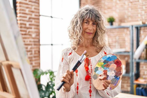 Женщина-художник средних лет уверенно улыбается, держа кисти и палитры в художественной студии - Фото, изображение